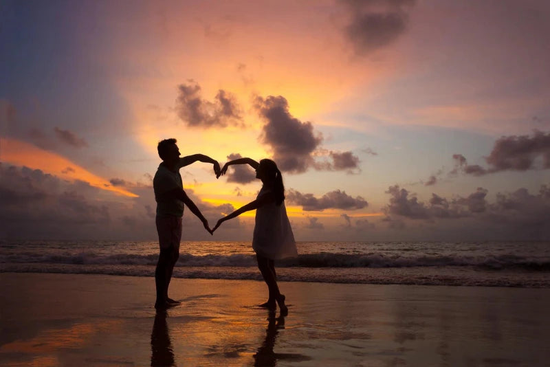 Two people standing by the ocean at sunset, making a heart with their arms. 